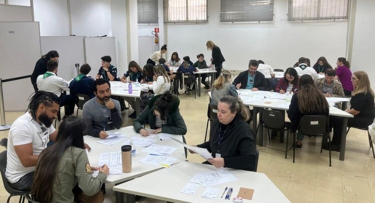 Pessoas reunidas em uma sala ampla preenchem cédulas juntas ao redor de diversas mesas brancas.