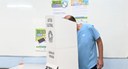 Foto de um homem vestindo camiseta azul de costas e levemente curvado para votar dentro de uma c...