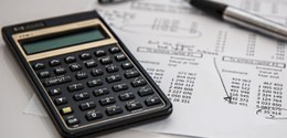foto de uma calculadora preta com detalhes dourados; ao fundo, uma folha de papel com números e ...