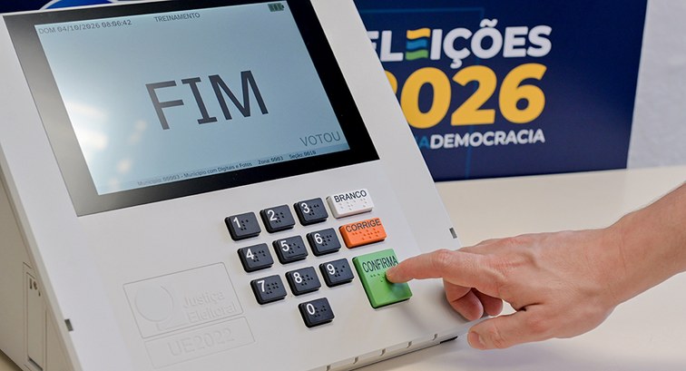 Uma imagem em plano médio exibe uma urna eletrônica branca sobre uma mesa cinza, ao lado de um b...