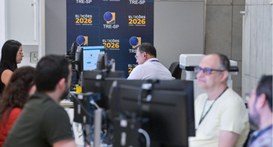 Pessoas trabalhando em bancadas de computadores no TRE-SP, com um painel azul ao fundo com a ins...