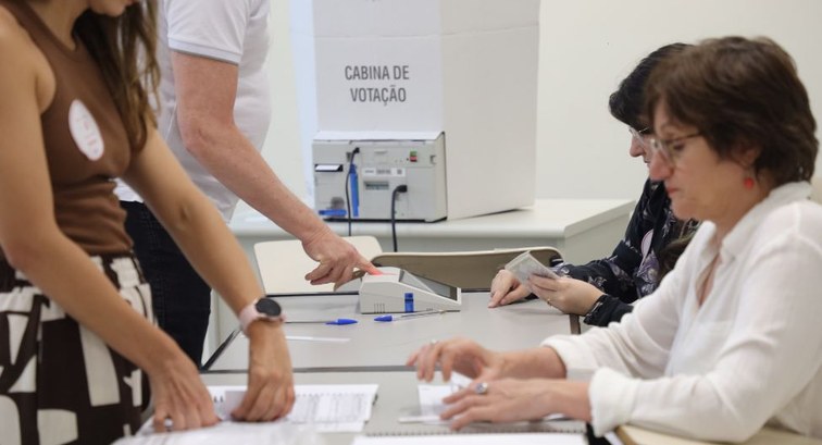 Eleitor posiciona o dedo no leitor biométrico sobre a mesa da seção eleitoral diante de mesárias...