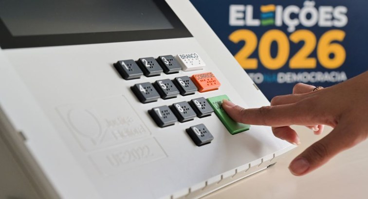 Fotografia em plano detalhe de uma urna eletrônica. A mão de uma pessoa, no canto direito da ima...